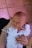 A newborn baby wrapped in a white garment is held gently by an adult's hands. The infant's eyes are slightly open, and there is a soft expression on their face. The background consists of a tiled floor with a mix of light and dark shades.