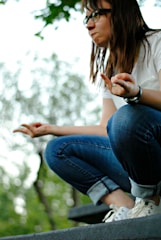 A person with long hair and glasses is crouching on a step, wearing a white shirt, rolled-up jeans, and sneakers. Their hands are in a meditative gesture with index fingers touching thumbs. The background is blurred with foliage and soft natural light.