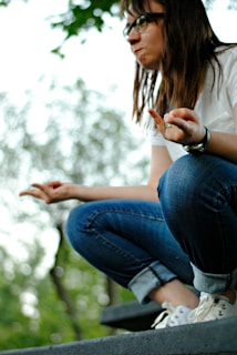 A person with long hair and glasses is crouching on a step, wearing a white shirt, rolled-up jeans, and sneakers. Their hands are in a meditative gesture with index fingers touching thumbs. The background is blurred with foliage and soft natural light.