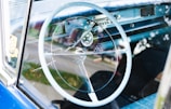 A classic car interior featuring a large white steering wheel, a blue dashboard, and vintage control dials. The radio and speedometer are visible, and there's an American flag hanging from the rearview mirror.