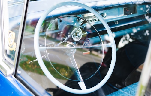 A classic car interior featuring a large white steering wheel, a blue dashboard, and vintage control dials. The radio and speedometer are visible, and there's an American flag hanging from the rearview mirror.