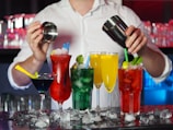 A bartender is preparing cocktails with various colorful drinks laid out in front. There are red, green, yellow, and blue drinks in distinct glasses, garnished with items like lime and mint. Ice cubes are scattered across the counter, adding to the refreshing vibe.