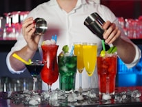 A bartender is preparing cocktails with various colorful drinks laid out in front. There are red, green, yellow, and blue drinks in distinct glasses, garnished with items like lime and mint. Ice cubes are scattered across the counter, adding to the refreshing vibe.