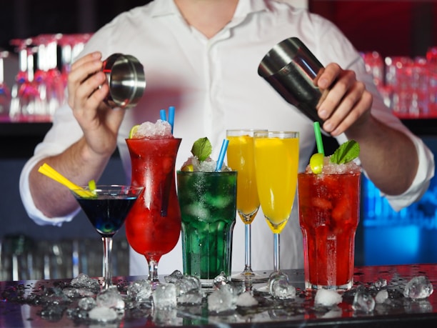 A bartender is preparing cocktails with various colorful drinks laid out in front. There are red, green, yellow, and blue drinks in distinct glasses, garnished with items like lime and mint. Ice cubes are scattered across the counter, adding to the refreshing vibe.