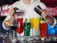 A bartender is preparing cocktails with various colorful drinks laid out in front. There are red, green, yellow, and blue drinks in distinct glasses, garnished with items like lime and mint. Ice cubes are scattered across the counter, adding to the refreshing vibe.