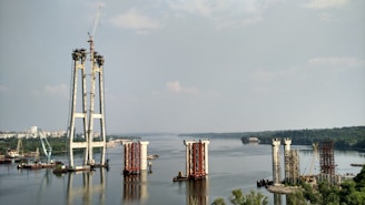 The image depicts a large-scale construction project in progress on a river. Several bridge pylons are partially built, with cranes and construction equipment visible around the site. The river extends into the distance, bordered by lush greenery and a cityscape.