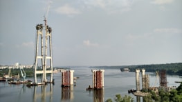 The image depicts a large-scale construction project in progress on a river. Several bridge pylons are partially built, with cranes and construction equipment visible around the site. The river extends into the distance, bordered by lush greenery and a cityscape.
