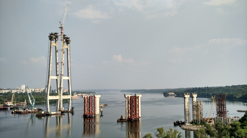 The image depicts a large-scale construction project in progress on a river. Several bridge pylons are partially built, with cranes and construction equipment visible around the site. The river extends into the distance, bordered by lush greenery and a cityscape.