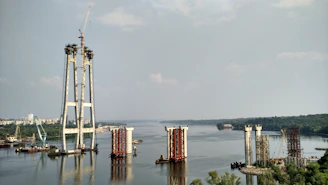 The image depicts a large-scale construction project in progress on a river. Several bridge pylons are partially built, with cranes and construction equipment visible around the site. The river extends into the distance, bordered by lush greenery and a cityscape.
