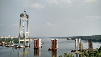 The image depicts a large-scale construction project in progress on a river. Several bridge pylons are partially built, with cranes and construction equipment visible around the site. The river extends into the distance, bordered by lush greenery and a cityscape.
