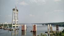 The image depicts a large-scale construction project in progress on a river. Several bridge pylons are partially built, with cranes and construction equipment visible around the site. The river extends into the distance, bordered by lush greenery and a cityscape.