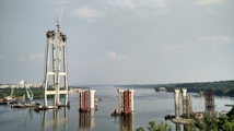 The image depicts a large-scale construction project in progress on a river. Several bridge pylons are partially built, with cranes and construction equipment visible around the site. The river extends into the distance, bordered by lush greenery and a cityscape.