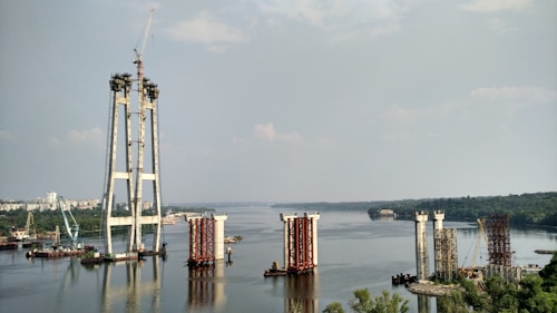 The image depicts a large-scale construction project in progress on a river. Several bridge pylons are partially built, with cranes and construction equipment visible around the site. The river extends into the distance, bordered by lush greenery and a cityscape.
