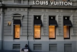 A luxury store facade featuring large windows illuminated from inside. Above the windows, the store's name is prominently displayed. The building's architecture is ornate, with detailed molding and classical accents. On the sidewalk, a person sits with a sign, creating a contrast between luxury and hardship.