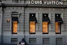 A luxury store facade featuring large windows illuminated from inside. Above the windows, the store's name is prominently displayed. The building's architecture is ornate, with detailed molding and classical accents. On the sidewalk, a person sits with a sign, creating a contrast between luxury and hardship.