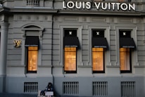 A luxury store facade featuring large windows illuminated from inside. Above the windows, the store's name is prominently displayed. The building's architecture is ornate, with detailed molding and classical accents. On the sidewalk, a person sits with a sign, creating a contrast between luxury and hardship.