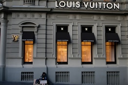 A luxury store facade featuring large windows illuminated from inside. Above the windows, the store's name is prominently displayed. The building's architecture is ornate, with detailed molding and classical accents. On the sidewalk, a person sits with a sign, creating a contrast between luxury and hardship.
