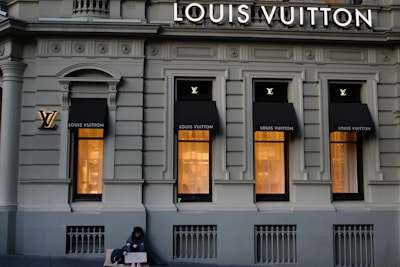 A luxury store facade featuring large windows illuminated from inside. Above the windows, the store's name is prominently displayed. The building's architecture is ornate, with detailed molding and classical accents. On the sidewalk, a person sits with a sign, creating a contrast between luxury and hardship.