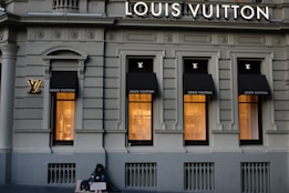 A luxury store facade featuring large windows illuminated from inside. Above the windows, the store's name is prominently displayed. The building's architecture is ornate, with detailed molding and classical accents. On the sidewalk, a person sits with a sign, creating a contrast between luxury and hardship.
