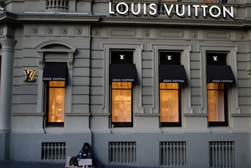 A luxury store facade featuring large windows illuminated from inside. Above the windows, the store's name is prominently displayed. The building's architecture is ornate, with detailed molding and classical accents. On the sidewalk, a person sits with a sign, creating a contrast between luxury and hardship.