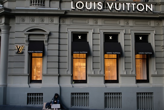 A luxury store facade featuring large windows illuminated from inside. Above the windows, the store's name is prominently displayed. The building's architecture is ornate, with detailed molding and classical accents. On the sidewalk, a person sits with a sign, creating a contrast between luxury and hardship.
