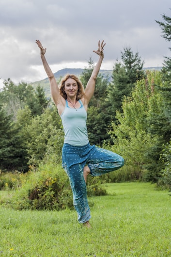 A person is practicing a yoga pose in a lush green outdoor setting. The individual is standing on one leg with arms raised to the sky, surrounded by trees and hills under a cloudy sky.