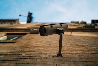 A surveillance camera is mounted on the side of a brick building, angled to observe the surroundings. The blue sky forms the background, and the image is taken from a low perspective looking upwards.