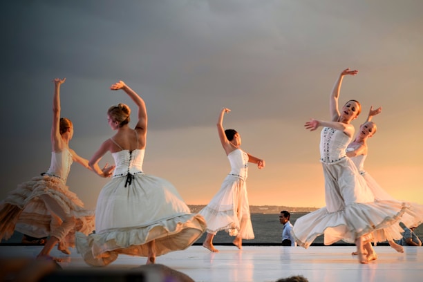 Several ballet dancers in flowing white dresses perform on stage with graceful movements. The scene is set against a dramatic sunset or evening sky, adding a warm glow to the performance. A few additional figures are visible in the background, seemingly watching or participating in the event.