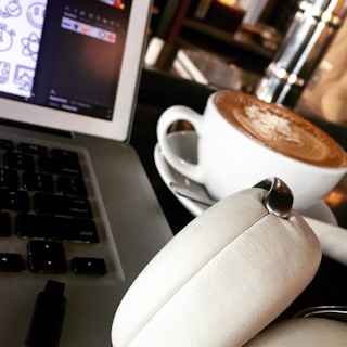 A close-up of a workspace featuring a laptop keyboard, a pair of white cushioned headphones, and a cup of coffee with latte art on a saucer. The headphones rest atop the laptop which shows an open design software interface. The setting hints at a cafe environment.