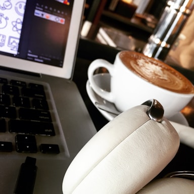 A close-up of a workspace featuring a laptop keyboard, a pair of white cushioned headphones, and a cup of coffee with latte art on a saucer. The headphones rest atop the laptop which shows an open design software interface. The setting hints at a cafe environment.