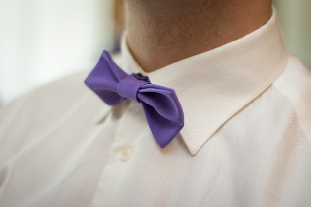 A close-up view of a person wearing a white dress shirt with a neatly tied purple bow tie. The shirt collar is crisp and well-ironed. The background appears soft and out of focus.
