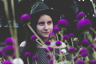 A woman wearing a dark green hat and patterned clothing is surrounded by vibrant purple flowers. The flowers are in the foreground, adding depth to the image, while the woman looks thoughtfully to the side.