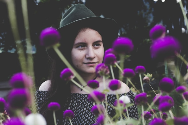 A woman wearing a dark green hat and patterned clothing is surrounded by vibrant purple flowers. The flowers are in the foreground, adding depth to the image, while the woman looks thoughtfully to the side.