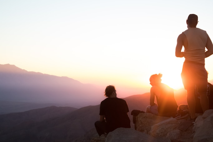 two people sitting and one man standing near cliff taken during golden hour in a national park