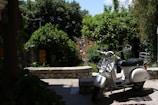 A white scooter is parked on a stone-paved area surrounded by lush greenery and trees. The background features a stone wall and a glimpse of buildings bathed in sunlight. There's a serene and quiet street ambiance with the scooter standing in partial shadow.