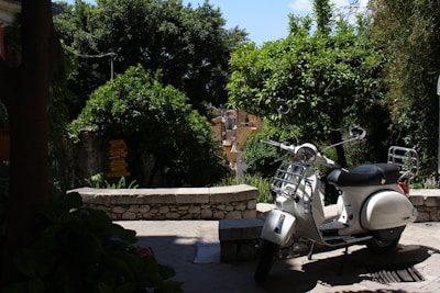 A white scooter is parked on a stone-paved area surrounded by lush greenery and trees. The background features a stone wall and a glimpse of buildings bathed in sunlight. There's a serene and quiet street ambiance with the scooter standing in partial shadow.