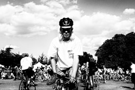 A black and white image capturing a group of cyclists gathered outdoors. A person in the foreground with a helmet and a t-shirt is prominently positioned, with more cyclists and trees visible in the background. The sky is partly cloudy.