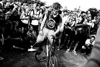 A crowd of people surrounds a cyclist who is positioned in the center. The cyclist is wearing a helmet and a t-shirt, holding onto the bicycle. Many in the crowd are smiling, taking photos, and appear to be enjoying the event. There is a sense of camaraderie and enthusiasm among the group.
