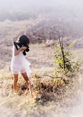 A young girl in a white dress is photographing a dry and sunny landscape with a camera. She is surrounded by dry grass and a leafless bush in a rural area, with a soft, natural lighting enhancing the serene setting.