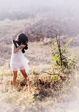 A young girl in a white dress is photographing a dry and sunny landscape with a camera. She is surrounded by dry grass and a leafless bush in a rural area, with a soft, natural lighting enhancing the serene setting.