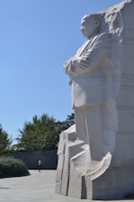 A large stone memorial sculpture of a man stands prominently against a clear blue sky. The figure is carved into a massive rock, with arms crossed and a solemn expression. Trees and a person are visible in the background, providing a sense of scale to the grand monument.