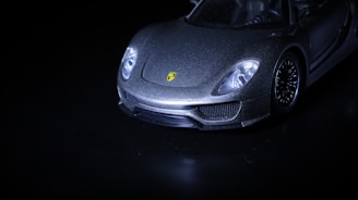 A sleek, luxury sports car is captured from a low-angle, highlighting its shining metallic body and distinctive logo on the front. The image is mostly dark, with the car emerging dramatically from the shadows.
