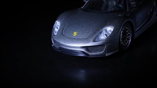 A sleek, luxury sports car is captured from a low-angle, highlighting its shining metallic body and distinctive logo on the front. The image is mostly dark, with the car emerging dramatically from the shadows.