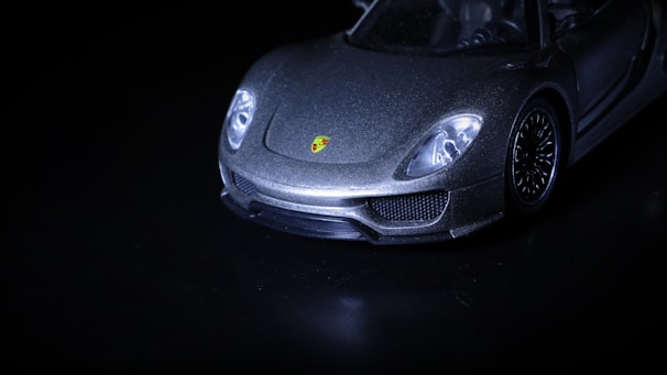 A sleek, luxury sports car is captured from a low-angle, highlighting its shining metallic body and distinctive logo on the front. The image is mostly dark, with the car emerging dramatically from the shadows.