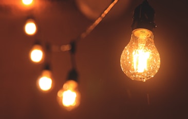 A series of illuminated vintage-style light bulbs hang against a dark background, casting a warm and inviting glow. The bulbs are arranged in a slightly diagonal line, with the largest bulb in focus showing visible filament details.