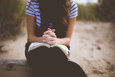 A person sits on a sandy beach with hands clasped in a prayer position over an open book. The background features blurred grass and sand, giving a peaceful and contemplative vibe.