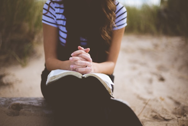 A person sits on a sandy beach with hands clasped in a prayer position over an open book. The background features blurred grass and sand, giving a peaceful and contemplative vibe.