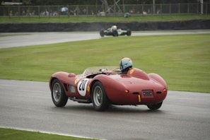 A vintage red racing car is being driven on a racetrack. The car has a white circular number 20 painted on the side and is equipped with a roll bar. The driver is wearing a blue helmet. In the background, another vintage car can be seen on the track, and there are spectators watching behind a barrier.