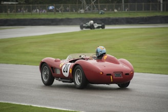 A vintage red racing car is being driven on a racetrack. The car has a white circular number 20 painted on the side and is equipped with a roll bar. The driver is wearing a blue helmet. In the background, another vintage car can be seen on the track, and there are spectators watching behind a barrier.