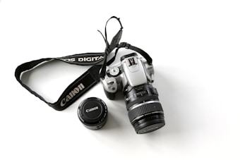 A silver digital camera with a detachable lens cap and a branded camera strap laid out on a white surface. The camera has various buttons and dials visible, suggesting a professional model.